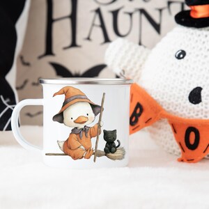May include: White enamel mug featuring a cartoon duck dressed as a witch, wearing an orange hat and holding a broom. A black cat sits on the broom. Halloween-themed design. The mug has a silver rim and a white handle.