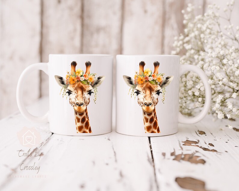 May include: Two white ceramic mugs with a giraffe wearing a flower crown printed on them. The giraffe has brown spots and a light brown face. The mugs are on a white wooden surface.