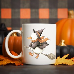 May include: White ceramic mug featuring a whimsical illustration of a duck dressed as a witch, riding a broom. The duck wears a black hat, coat, and orange scarf. The background includes pumpkins and fall leaves, perfect for Halloween.
