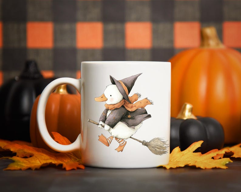 May include: White ceramic mug featuring a whimsical illustration of a duck dressed as a witch, riding a broom. The duck wears a black hat, coat, and orange scarf. The background includes pumpkins and fall leaves, perfect for Halloween.