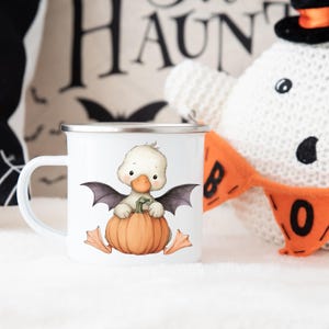 May include: White enamel mug with a Halloween-themed design. The mug features a cartoon duckling with bat wings, sitting on a pumpkin. The mug has a silver rim and handle. A Halloween-themed decorative pillow and a stuffed ghost are in the background.
