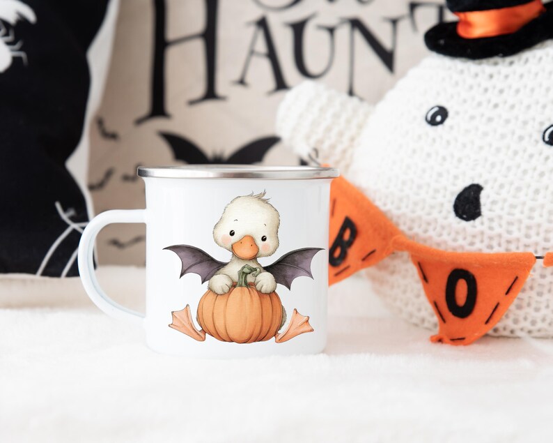 May include: White enamel mug with a Halloween-themed design. The mug features a cartoon duckling with bat wings, sitting on a pumpkin. The mug has a silver rim and handle. A Halloween-themed decorative pillow and a stuffed ghost are in the background.