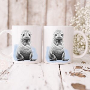 K&ouml;nnte beinhalten: Zwei wei&szlig;e Keramikbecher mit einer Schwarzwei&szlig;illustration eines Seehunds auf jedem. Der Seehund sitzt auf einem hellblauen Hintergrund.