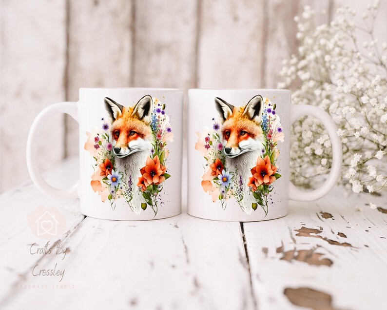 May include: Two white ceramic mugs with a watercolor design of a fox surrounded by flowers. The fox is orange and white with a black nose and eyes. The flowers are in shades of pink, purple, blue, and orange.