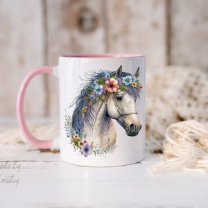 Pu&ograve; includere: Tazza in ceramica bianca con bordo e manico rosa. La tazza presenta un'illustrazione ad acquerello di un cavallo bianco con una corona di fiori. I fiori sono rosa, blu e viola.