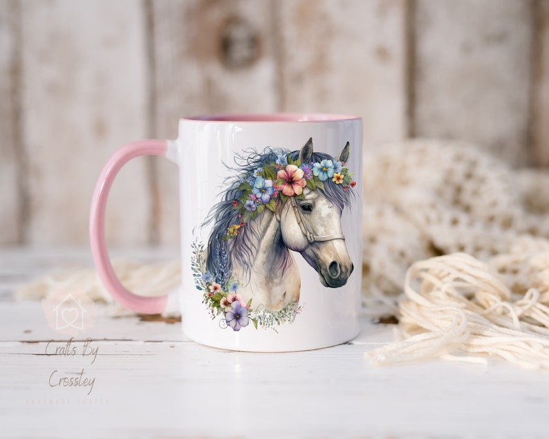 Pu&ograve; includere: Tazza in ceramica bianca con bordo e manico rosa. La tazza presenta un'illustrazione ad acquerello di un cavallo bianco con una corona di fiori. I fiori sono rosa, blu e viola.