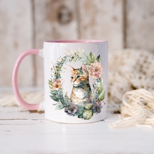 May include: A white ceramic mug with a pink rim and handle. The mug features a watercolor illustration of a cat surrounded by flowers and greenery.