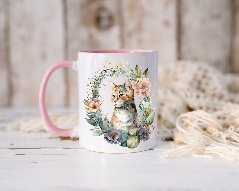 May include: A white ceramic mug with a pink rim and handle. The mug features a watercolor illustration of a cat surrounded by flowers and greenery.