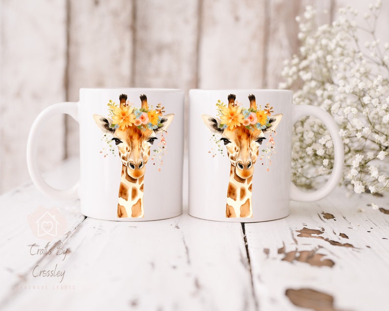 May include: Two white ceramic mugs with a watercolor design of a giraffe wearing a floral crown. The giraffe has brown spots and a light brown face. The flowers are yellow, orange, and blue.
