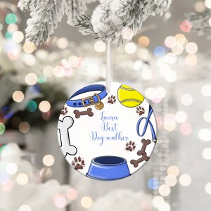 May include: Round Christmas ornament with a dog theme. The ornament features a blue dog collar, tennis ball, paw prints, and a dog bowl. The text "Laura Best Dog Walker" is written in blue script. The ornament is white with a ribbon for hanging.