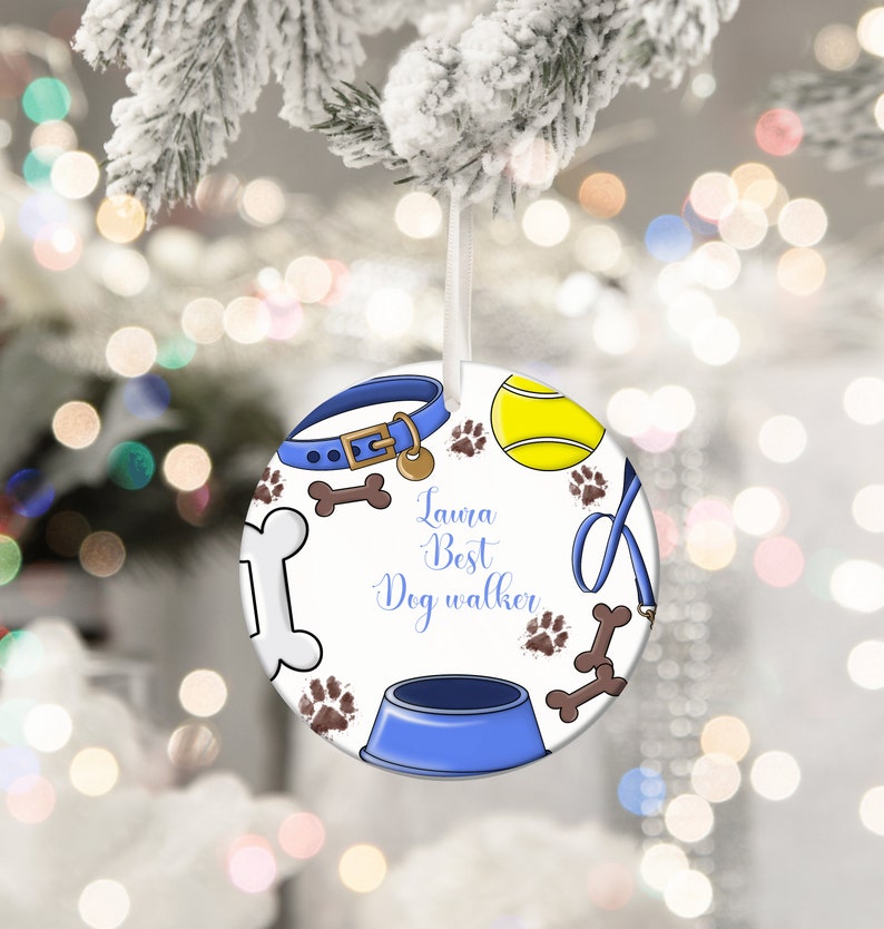 May include: Round Christmas ornament with a dog theme. The ornament features a blue dog collar, tennis ball, paw prints, and a dog bowl. The text "Laura Best Dog Walker" is written in blue script. The ornament is white with a ribbon for hanging.