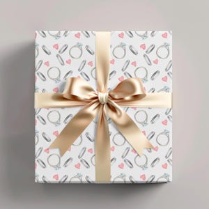 May include: A wrapped gift box with a white background featuring a pattern of diamond rings and pink hearts. A gold satin ribbon and bow are tied around the box, creating a festive presentation.