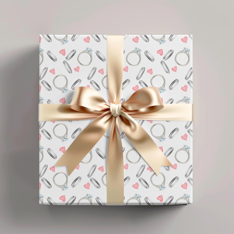 May include: A wrapped gift box with a white background featuring a pattern of diamond rings and pink hearts. A gold satin ribbon and bow are tied around the box, creating a festive presentation.