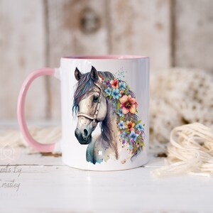 Pu&ograve; includere: Tazza in ceramica bianca con bordo e manico rosa. La tazza presenta un'illustrazione ad acquerello di un cavallo bianco con una corona di fiori rosa, blu e gialli.