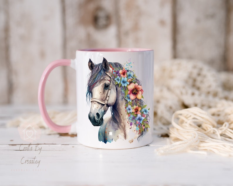 Pu&ograve; includere: Tazza in ceramica bianca con bordo e manico rosa. La tazza presenta un'illustrazione ad acquerello di un cavallo bianco con una corona di fiori rosa, blu e gialli.