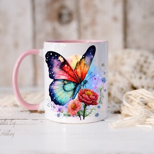 K&ouml;nnte beinhalten: Eine wei&szlig;e Keramiktasse mit einem rosa Henkel zeigt einen bunten Schmetterling mit galaxieartigen Fl&uuml;geln und einem Aquarellhintergrund. Der Schmetterling ist von rosa, blauen und gr&uuml;nen Blumen umgeben.