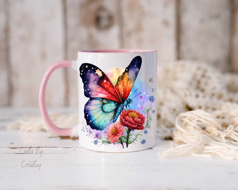 K&ouml;nnte beinhalten: Eine wei&szlig;e Keramiktasse mit einem rosa Henkel zeigt einen bunten Schmetterling mit galaxieartigen Fl&uuml;geln und einem Aquarellhintergrund. Der Schmetterling ist von rosa, blauen und gr&uuml;nen Blumen umgeben.