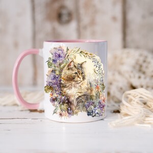 May include: A white ceramic mug with a pink rim features a watercolor illustration of a cat sitting in a bed of purple and blue flowers. The cat has brown and white fur and green eyes.