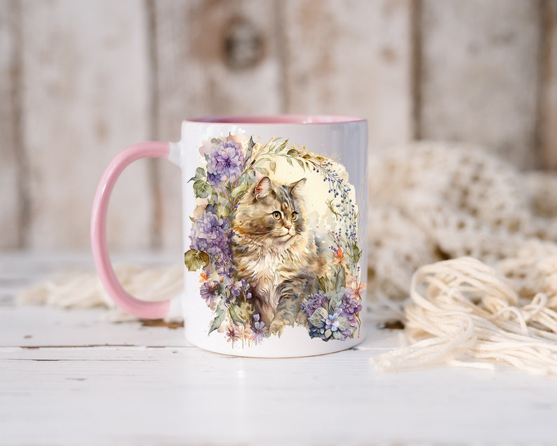 May include: A white ceramic mug with a pink rim features a watercolor illustration of a cat sitting in a bed of purple and blue flowers. The cat has brown and white fur and green eyes.
