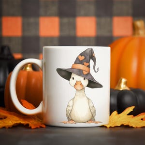 May include: White ceramic mug featuring a watercolor illustration of a duck wearing a witch hat with orange heart accents. The background includes pumpkins and fall leaves, perfect for Halloween.