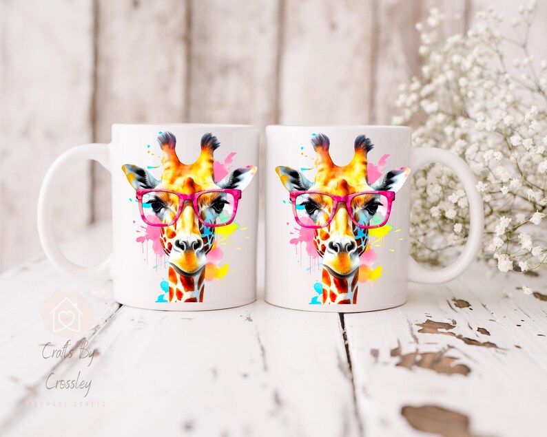 May include: Two white ceramic mugs with a colorful watercolor giraffe wearing pink glasses. The giraffe is surrounded by colorful paint splatters.