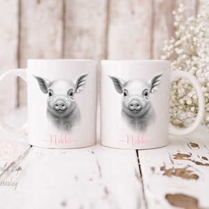 May include: Two white ceramic mugs featuring a black and white illustration of a piglet on each. The mugs are personalised with the name "Nikkie" written in pink at the bottom.