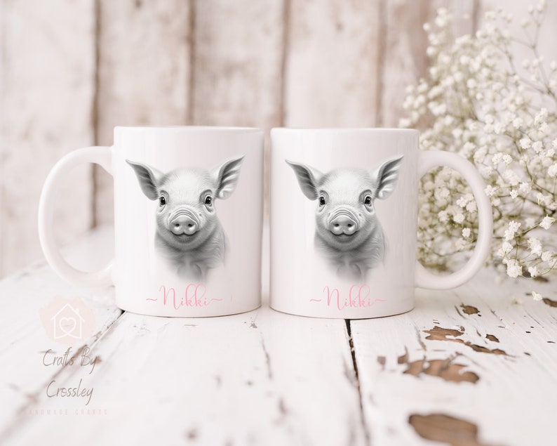 May include: Two white ceramic mugs featuring a black and white illustration of a piglet on each. The mugs are personalised with the name "Nikkie" written in pink at the bottom.