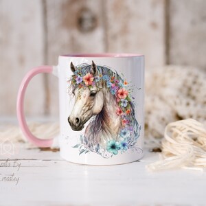 Pu&ograve; includere: Tazza in ceramica bianca con bordo e manico rosa. La tazza presenta un'illustrazione ad acquerello di un cavallo bianco con una corona di fiori. I fiori sono rosa, blu e viola.