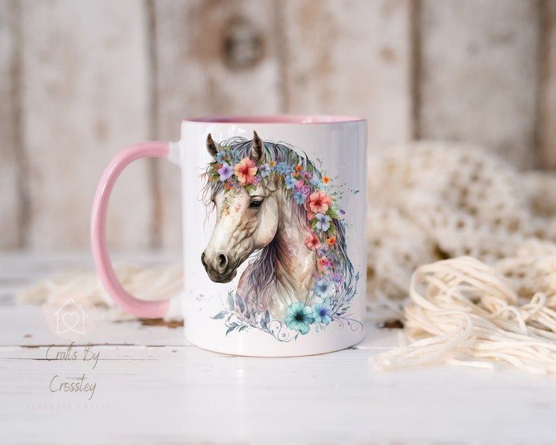 Pu&ograve; includere: Tazza in ceramica bianca con bordo e manico rosa. La tazza presenta un'illustrazione ad acquerello di un cavallo bianco con una corona di fiori. I fiori sono rosa, blu e viola.