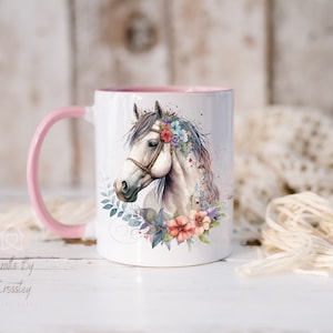 Pu&ograve; includere: Una tazza in ceramica bianca con un manico e un bordo rosa. La tazza presenta un'illustrazione ad acquerello di un cavallo bianco con una corona di fiori. La testa del cavallo &egrave; girata di lato e i suoi occhi sono chiusi. I fiori della corona sono rosa, blu e viola.