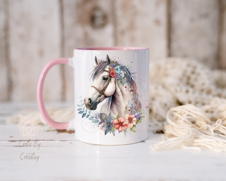 Pu&ograve; includere: Una tazza in ceramica bianca con un manico e un bordo rosa. La tazza presenta un'illustrazione ad acquerello di un cavallo bianco con una corona di fiori. La testa del cavallo &egrave; girata di lato e i suoi occhi sono chiusi. I fiori della corona sono rosa, blu e viola.