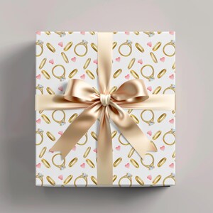May include: A wrapped gift box with a white background featuring a pattern of gold rings and pink hearts. A gold satin ribbon and bow are tied around the box. The bow is centered on the top of the box.