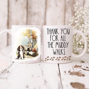 May include: Two white ceramic mugs. One mug features a watercolor illustration of a dog sitting near logs and trees. The other mug has the text "THANK YOU FOR ALL THE MUDDY WALKS" with paw prints.