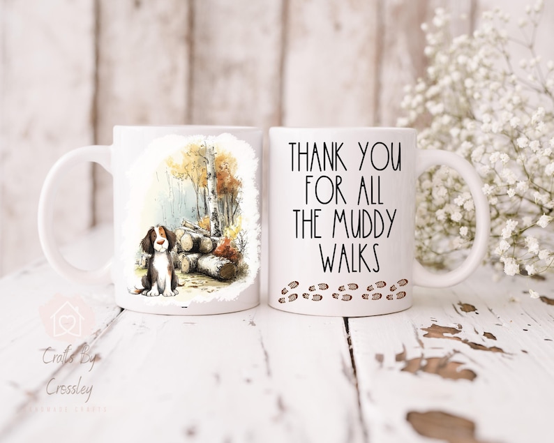 May include: Two white ceramic mugs. One mug features a watercolor illustration of a dog sitting near logs and trees. The other mug has the text "THANK YOU FOR ALL THE MUDDY WALKS" with paw prints.