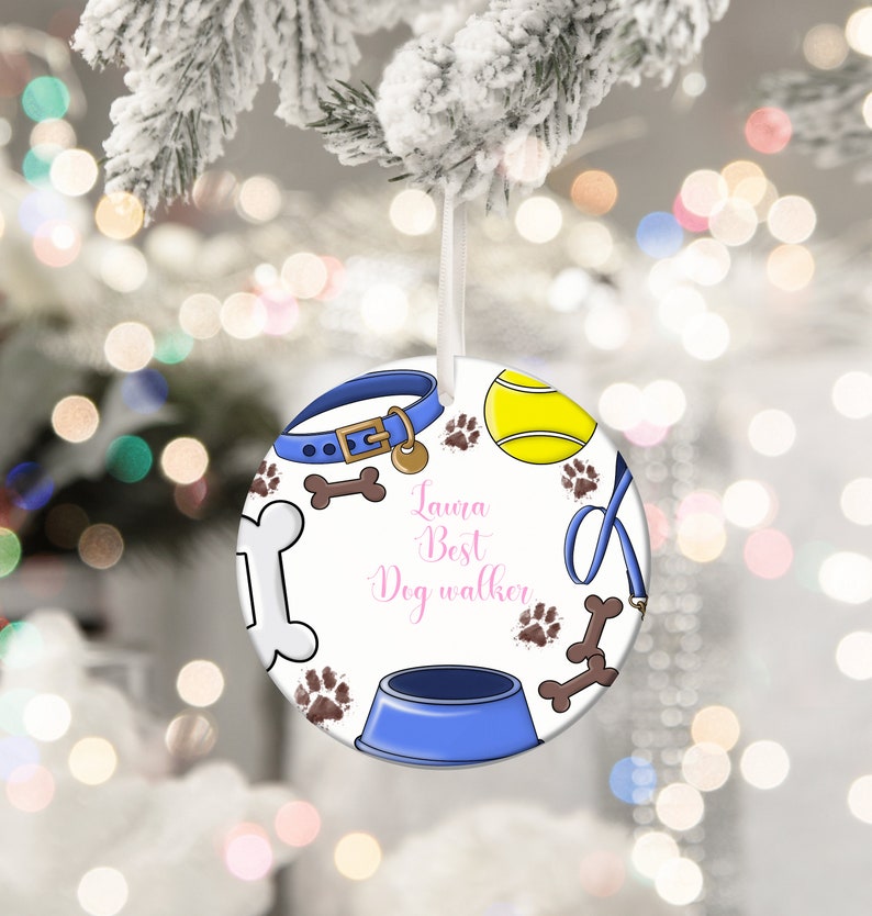 May include: Round white Christmas ornament with a blue dog collar, tennis ball, paw prints, and dog bone illustrations. The ornament has a blue dog bowl and the text "Laura Best Dog walker" in pink script.