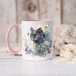 May include: A white ceramic mug with a pink rim and handle features a watercolor illustration of a gray cat with green eyes surrounded by flowers and butterflies.