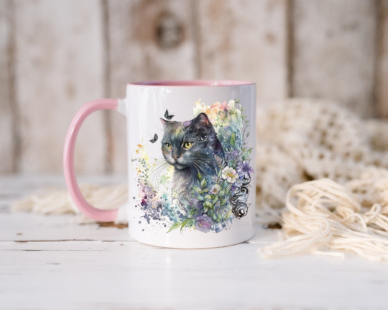 May include: A white ceramic mug with a pink rim and handle features a watercolor illustration of a gray cat with green eyes surrounded by flowers and butterflies.