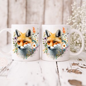 May include: Two white ceramic mugs with a fox head design. The fox is surrounded by watercolor flowers in shades of pink, purple, and yellow. The mugs are on a white wooden surface.