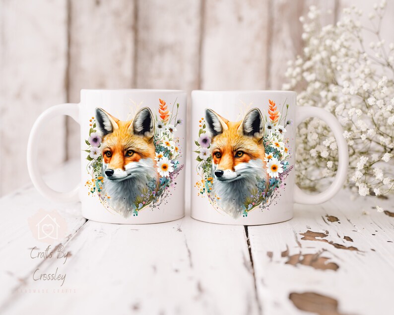 May include: Two white ceramic mugs with a fox head design. The fox is surrounded by watercolor flowers in shades of pink, purple, and yellow. The mugs are on a white wooden surface.