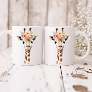 May include: Two white ceramic mugs with a watercolor illustration of a giraffe wearing a floral crown. The giraffe has brown spots and a light brown face. The mugs are on a white wooden surface.