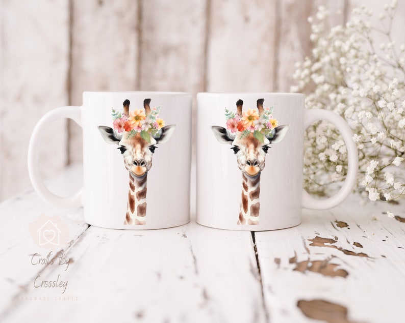 May include: Two white ceramic mugs with a watercolor illustration of a giraffe wearing a floral crown. The giraffe has brown spots and a light brown face. The mugs are on a white wooden surface.
