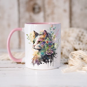 May include: A white ceramic mug with a pink handle and a watercolor illustration of a cat with flowers on its head. The cat is looking to the right and has green eyes.