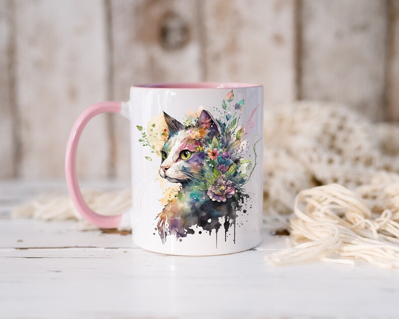 May include: A white ceramic mug with a pink handle and a watercolor illustration of a cat with flowers on its head. The cat is looking to the right and has green eyes.