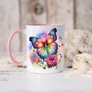 K&ouml;nnte beinhalten: Eine wei&szlig;e Keramiktasse mit pinkfarbenem Henkel zeigt einen bunten Schmetterling mit Galaxie-Design und Aquarellblumen. Die Tasse steht auf einer wei&szlig;en Holzoberfl&auml;che.