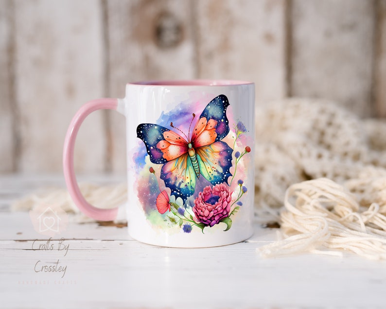 K&ouml;nnte beinhalten: Eine wei&szlig;e Keramiktasse mit pinkfarbenem Henkel zeigt einen bunten Schmetterling mit Galaxie-Design und Aquarellblumen. Die Tasse steht auf einer wei&szlig;en Holzoberfl&auml;che.