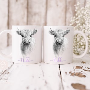 May include: Two white ceramic mugs featuring black and white illustrations of a Highland cow. The mugs are personalised with the name "Nikki" in purple at the bottom.