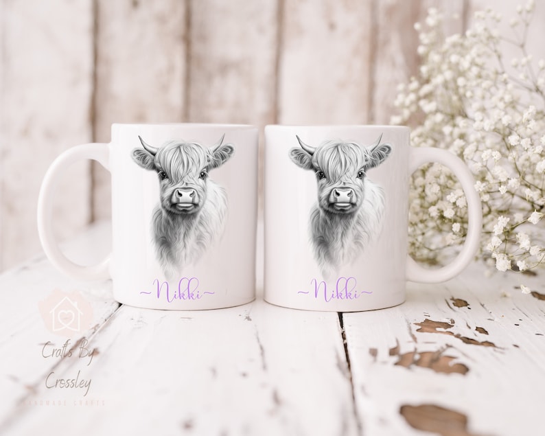 May include: Two white ceramic mugs featuring black and white illustrations of a Highland cow. The mugs are personalised with the name "Nikki" in purple at the bottom.