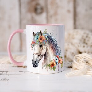 Pu&ograve; includere: Tazza in ceramica bianca con manico rosa e un'illustrazione ad acquerello di un cavallo bianco con una corona di fiori. La testa del cavallo &egrave; rivolta verso il lato sinistro della tazza. I fiori della corona sono rosa, blu e arancioni.