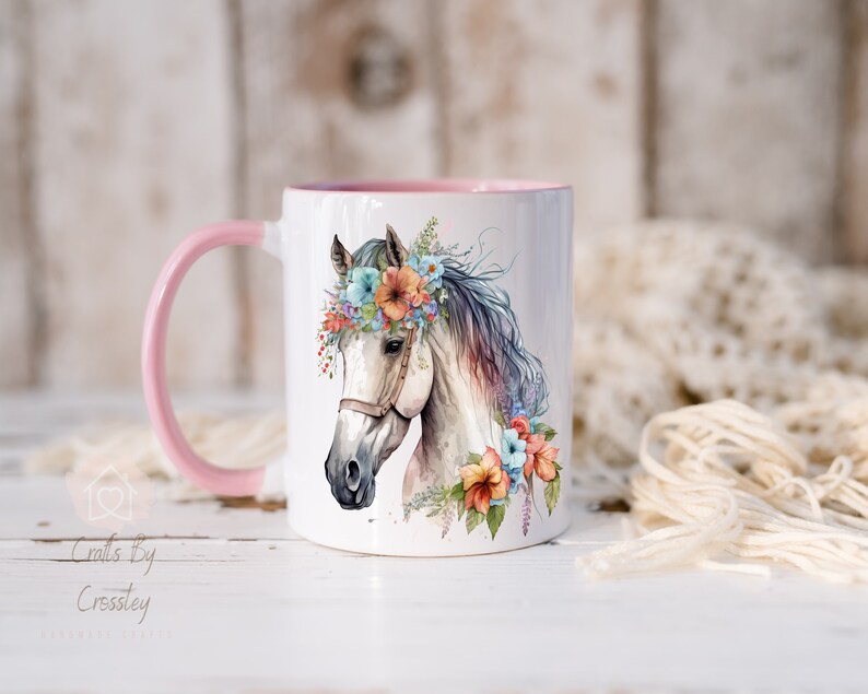 Pu&ograve; includere: Tazza in ceramica bianca con manico rosa e un'illustrazione ad acquerello di un cavallo bianco con una corona di fiori. La testa del cavallo &egrave; rivolta verso il lato sinistro della tazza. I fiori della corona sono rosa, blu e arancioni.
