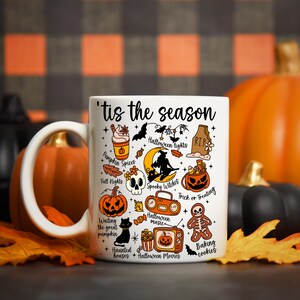 Pu&ograve; includere: Tazza in ceramica bianca con la scritta "'tis the season" e illustrazioni a tema Halloween. La tazza presenta immagini di zucche, pipistrelli, una strega e altri elementi spettrali. La tazza &egrave; circondata da zucche e foglie autunnali.